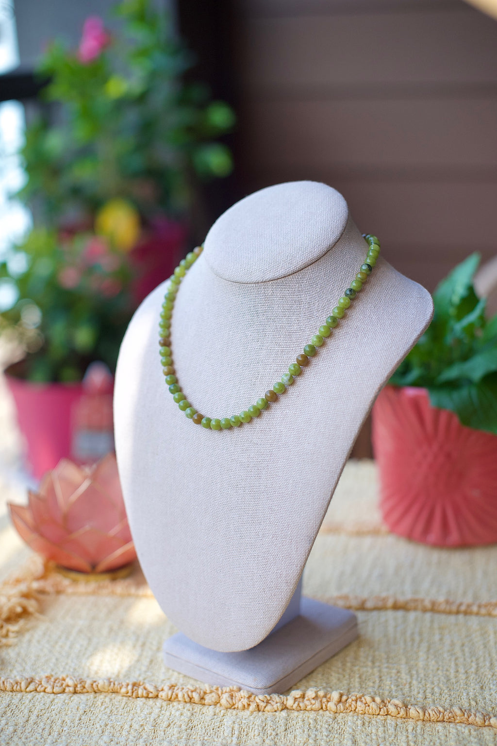 A green beaded necklace displayed on a light beige fabric jewelry bust, set against a softly blurred background with potted plants and warm indoor lighting.