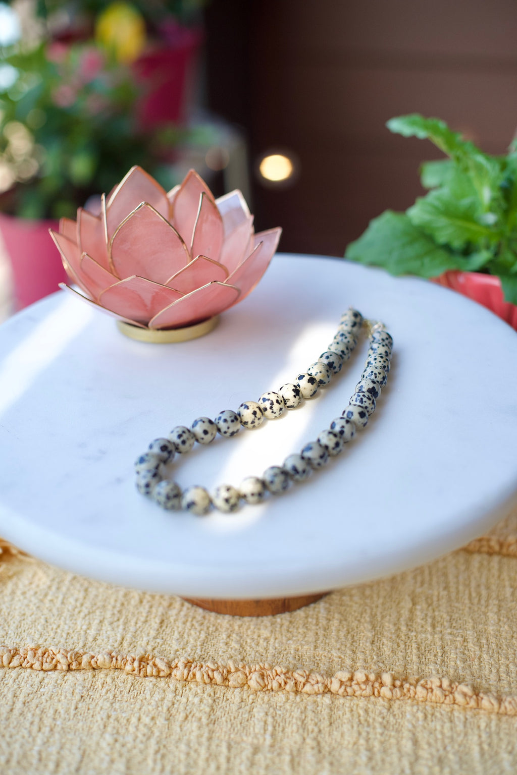 A beaded bracelet with speckled white, black, and gray beads arranged on a round white marble tray, styled with a pink lotus candle holder and green plants in the background.
