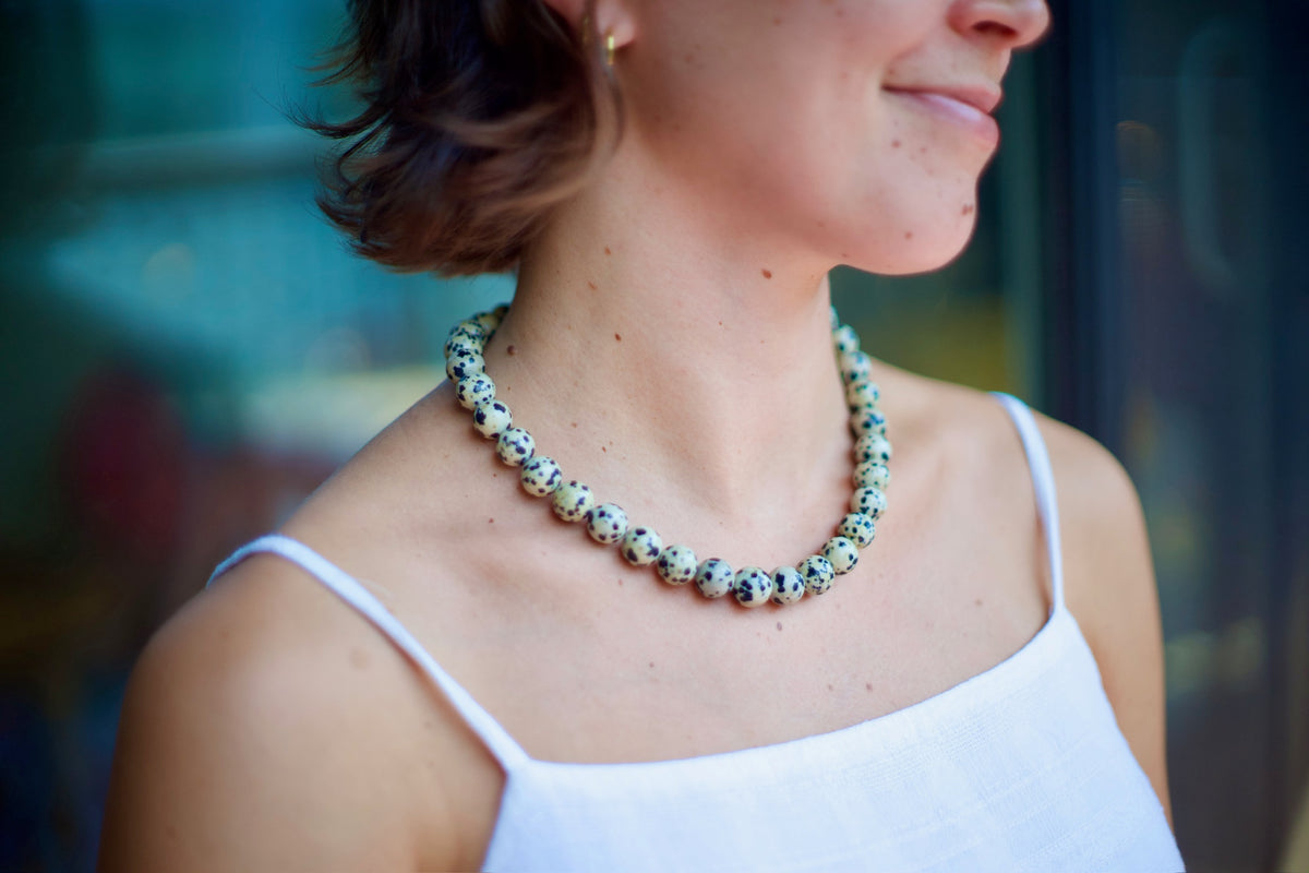 A close-up side profile of a woman wearing a beaded necklace made of speckled white beads with black and blue flecks, paired with a white spaghetti-strap top.
