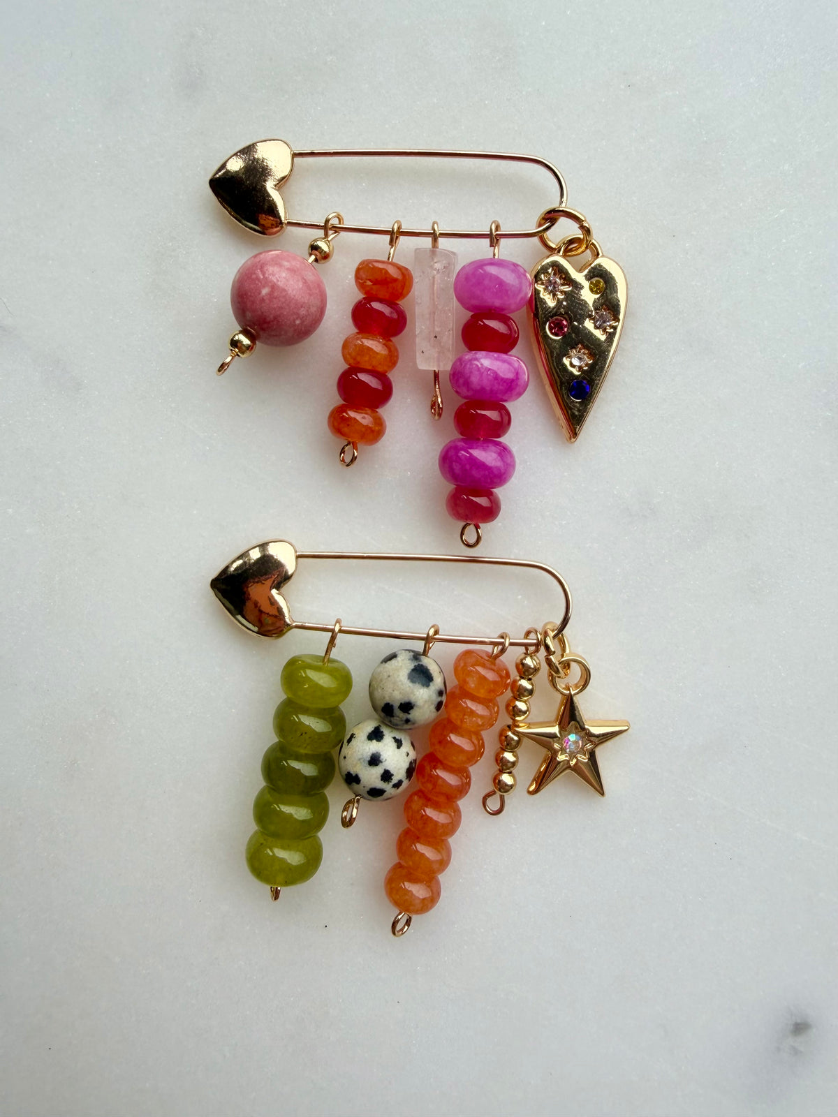 Two gold safety-pin style bag charms with dangling colorful beads and charms, displayed on a light marble surface. The top bag charm features pink, orange, and magenta beads with a heart charm, and the bottom bag charm features olive green, orange, and black-and-white spotted beads with a star charm.
