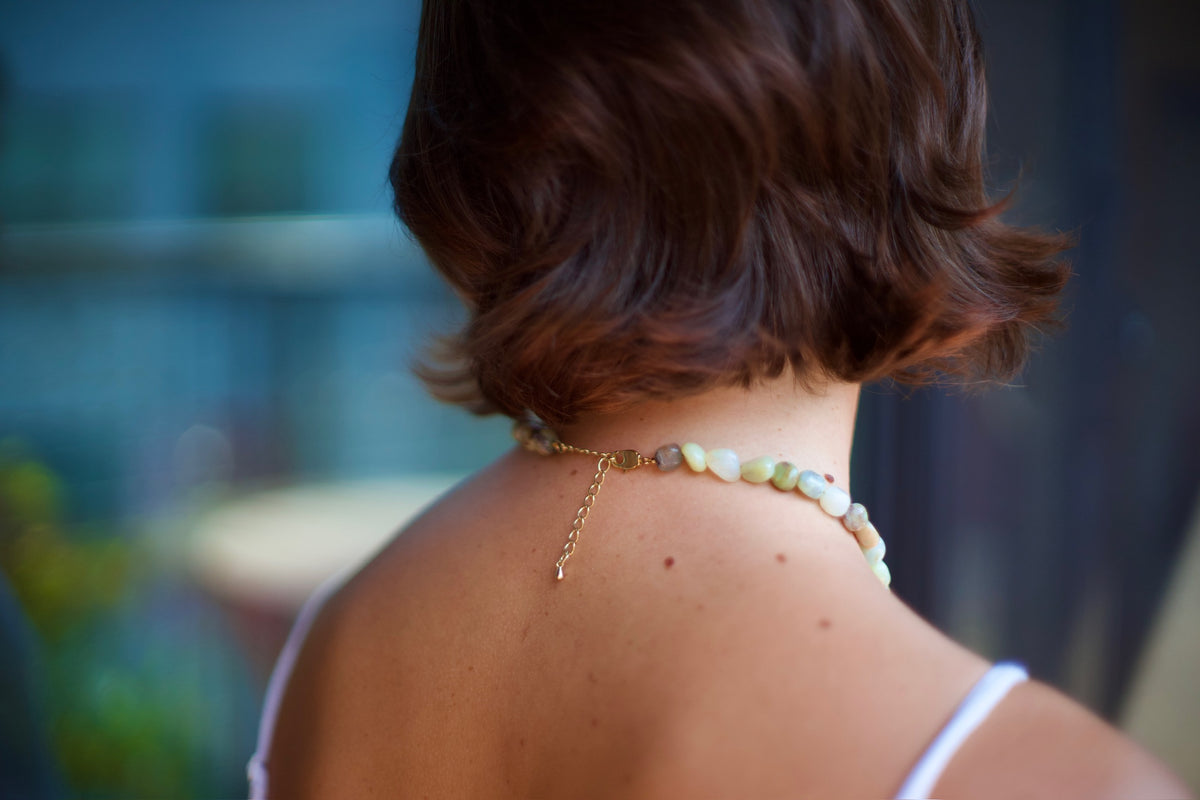 A close-up back view of a person with short brown hair wearing a delicate beaded necklace with a gold chain extender, set against a softly blurred blue-toned background.