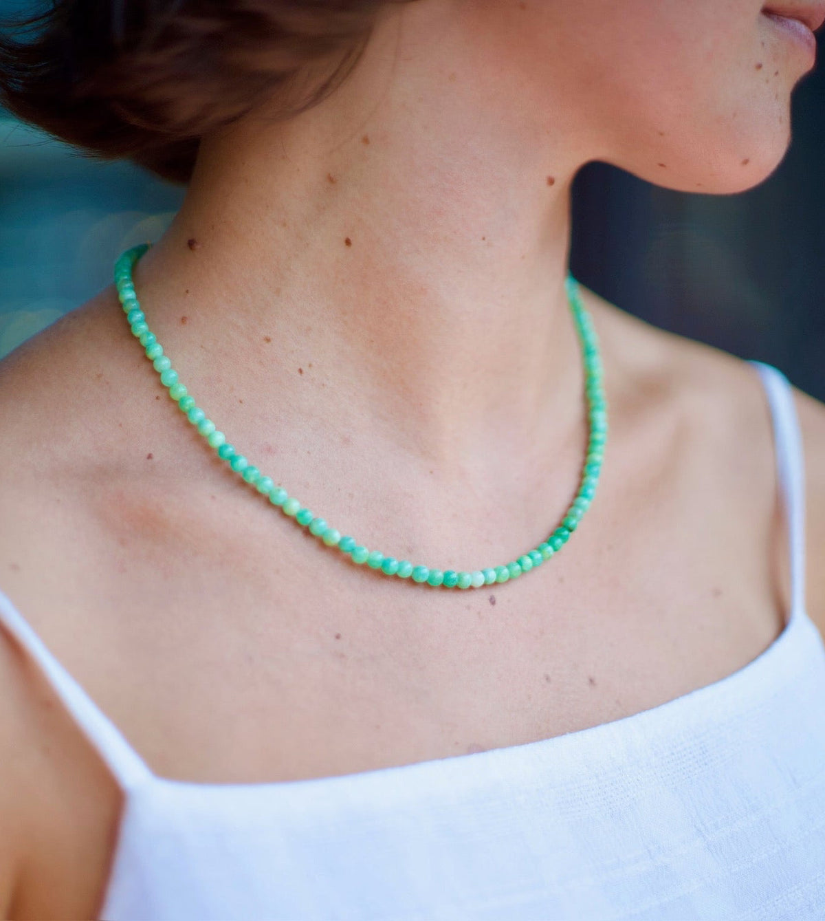 A close-up lifestyle shot of a woman wearing a delicate mint green beaded necklace with a white spaghetti-strap top, shown from the side.