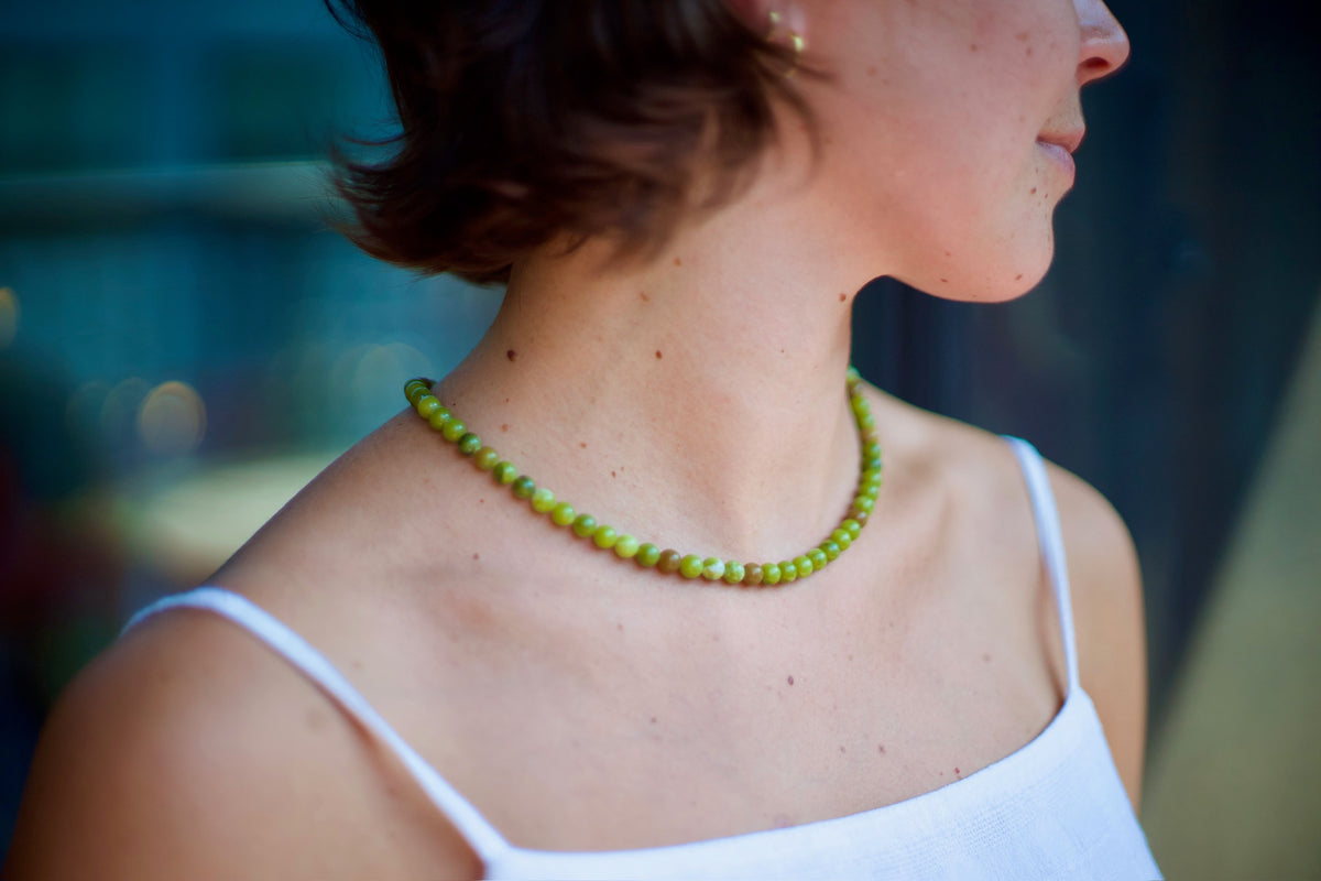 A close-up side profile of a woman wearing a chunky lime green beaded necklace and a white spaghetti-strap top, set against a blurred dark background.