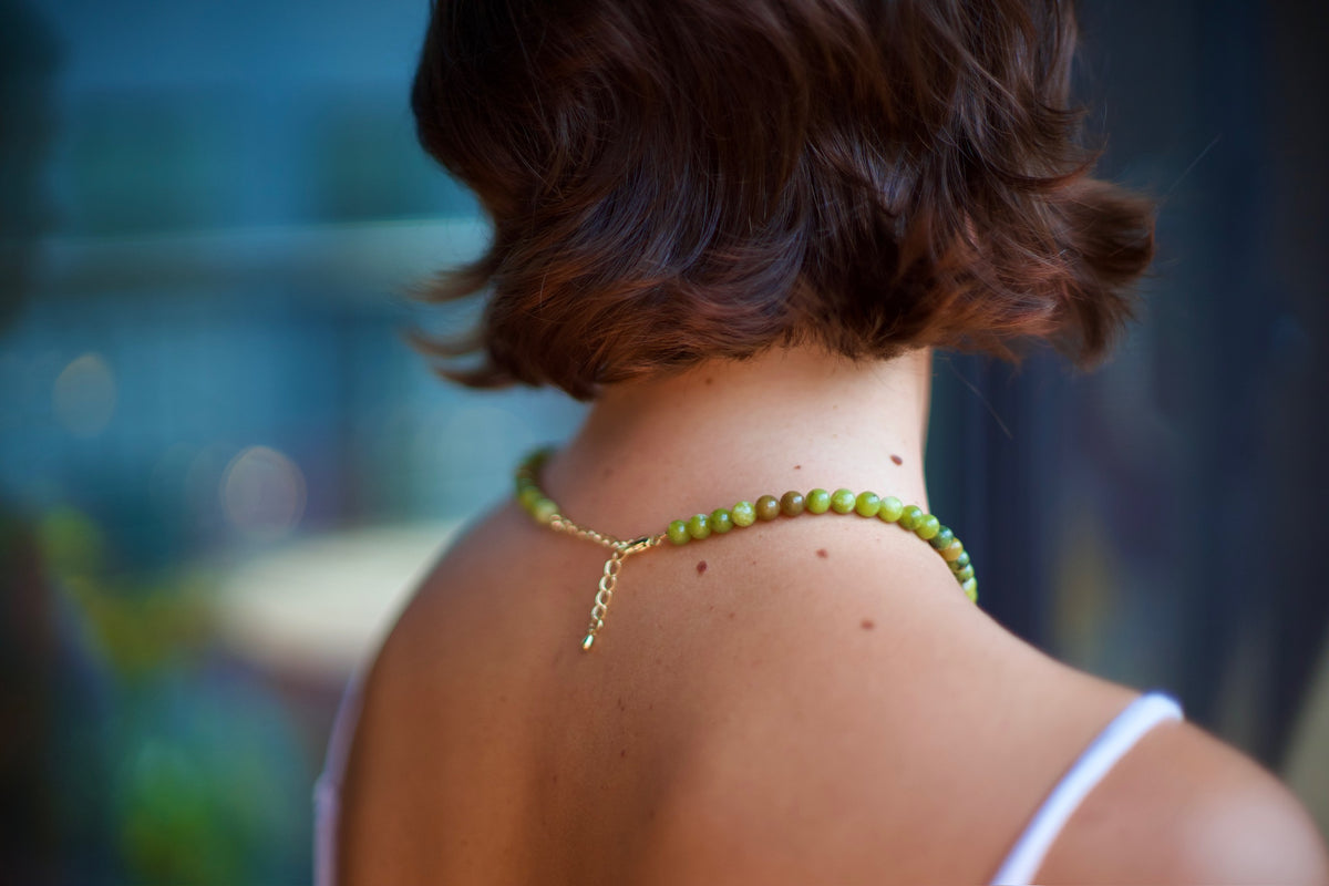 A close-up back view of a woman with short brown hair wearing a green beaded necklace with a gold clasp and a small dangling charm.