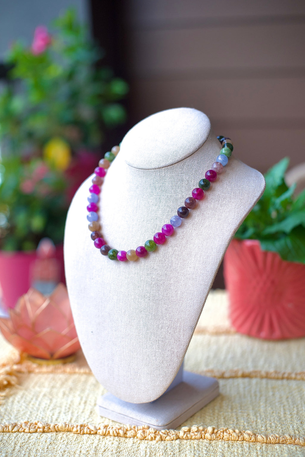 A multicolor beaded necklace displayed on a cream fabric jewelry bust, with a softly blurred background of potted plants and a wooden wall.