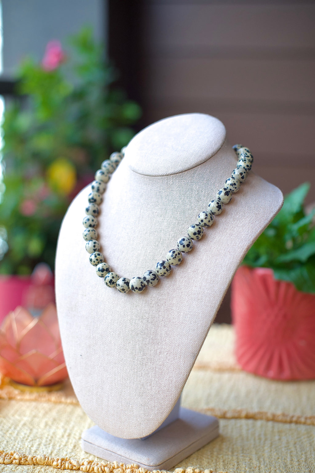 A beaded necklace with speckled gray, black, and white beads displayed on a cream-colored fabric jewelry bust. The background is softly blurred with green plants and pink decorative elements.