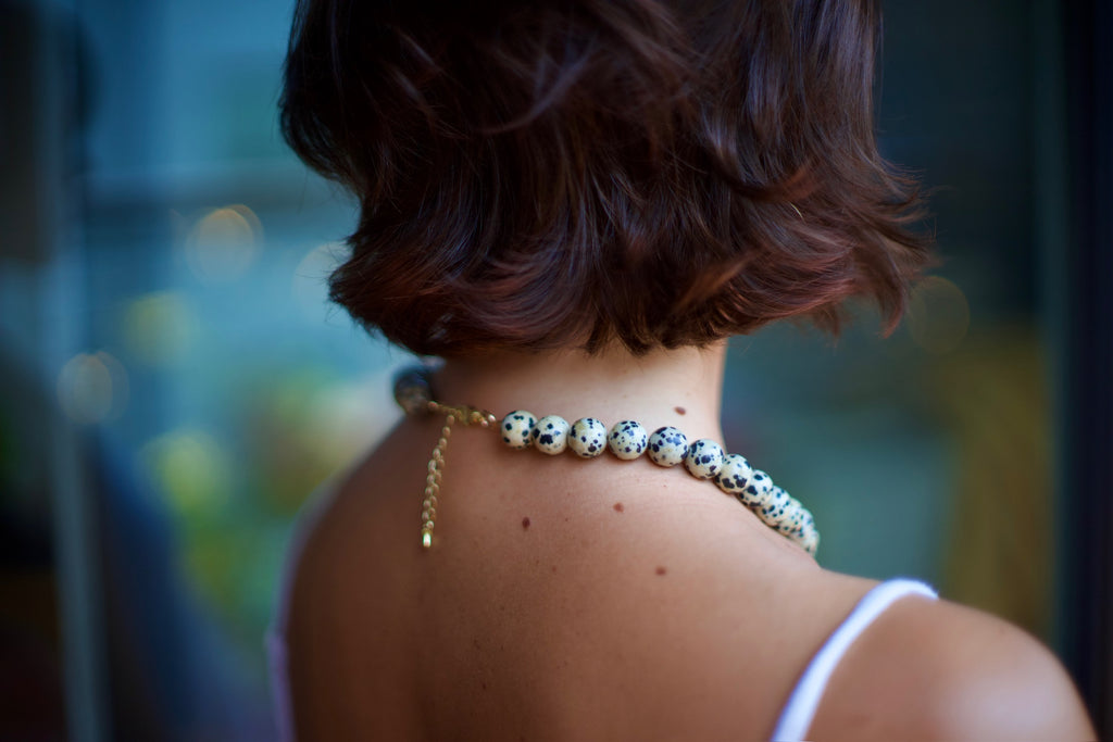 A close-up back view of a woman with short brown hair wearing a beaded choker necklace with a gold chain and clasp.