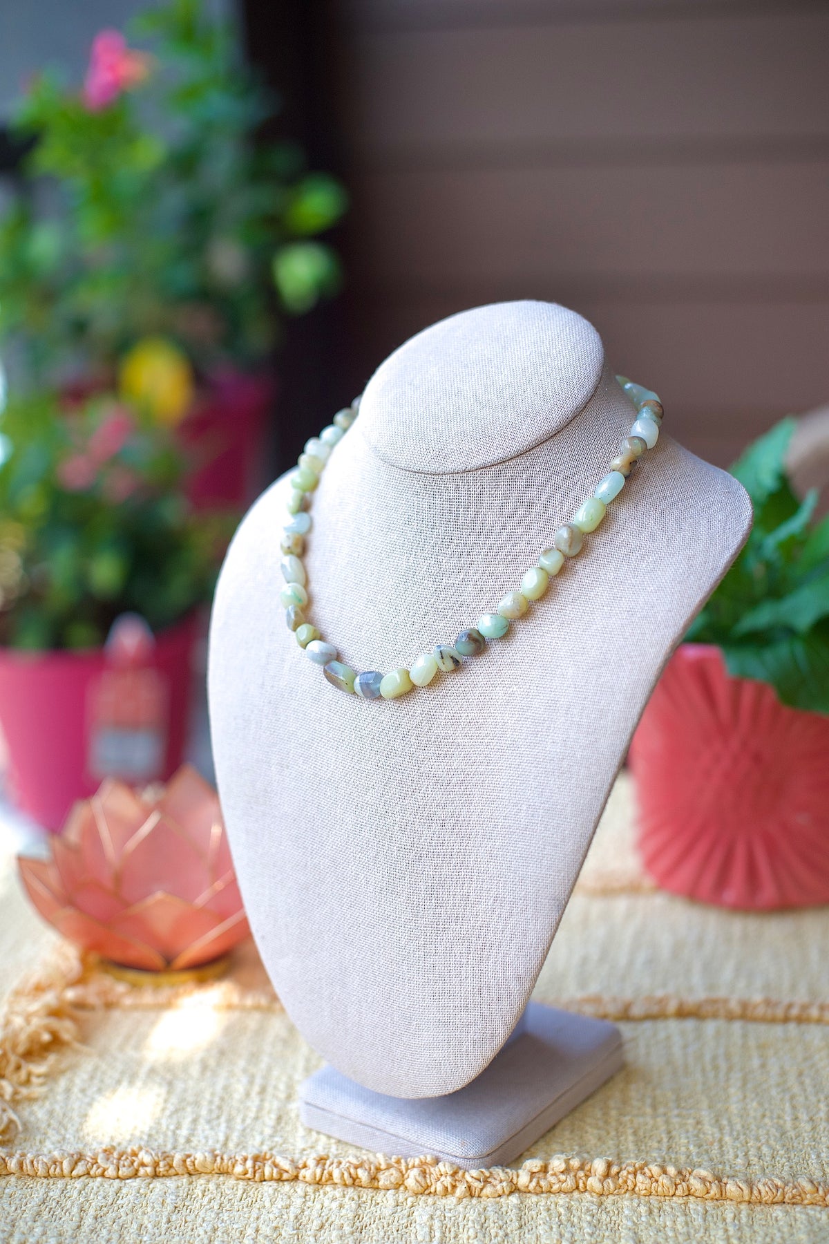 A beaded necklace made of irregular, polished stones in soft pastel shades of green, blue, and cream, displayed on a light beige fabric jewelry bust. The background is a warm, softly blurred setting with potted plants and a textured table runner.