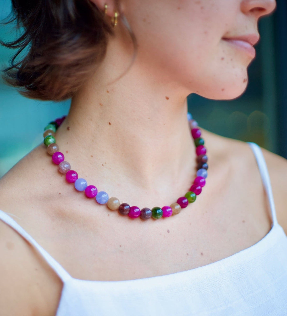 A close-up lifestyle shot of a woman wearing a colorful beaded necklace. The necklace features a mix of vibrant and pastel beads including pink, purple, green, blue, and brown.