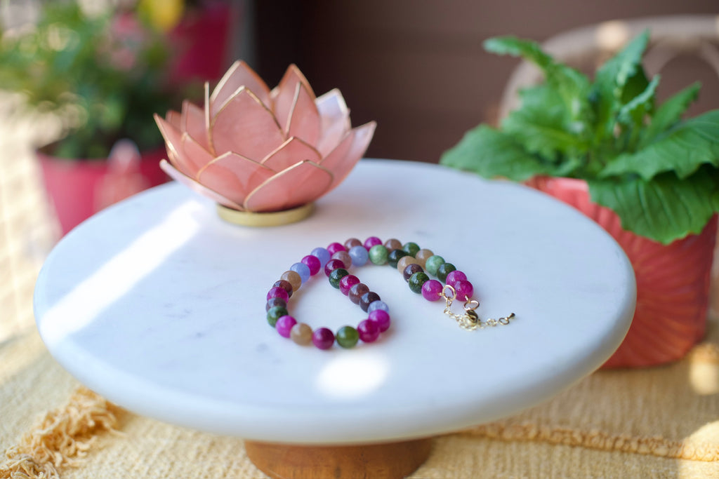 A beaded gemstone necklace with purple, green, and pink round beads and a gold clasp, displayed on a white marble pedestal with a pink lotus flower and potted plants in the background.