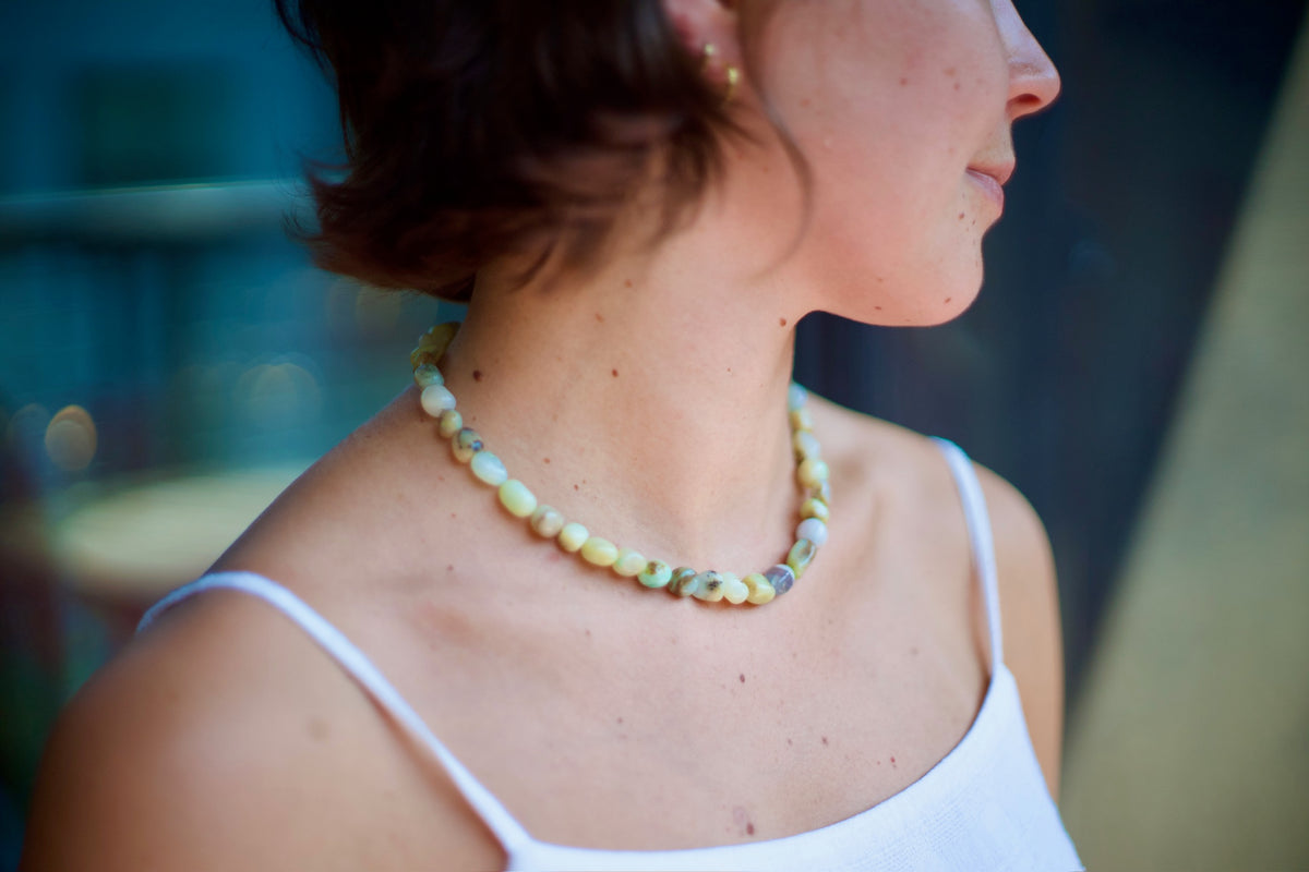 A close-up side profile of a woman wearing a chunky beaded necklace made of natural marbled stone beads in muted yellow, green, and gray tones, paired with a white spaghetti-strap top.