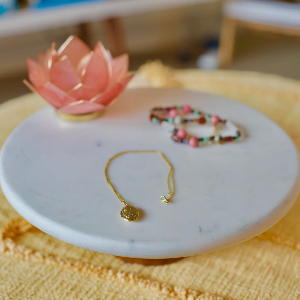 A gold-toned delicate bracelet with a small round disc charm and a star accent, displayed on a round white marble tray next to a pink lotus flower and a beaded bracelet on a yellow textured blanket.