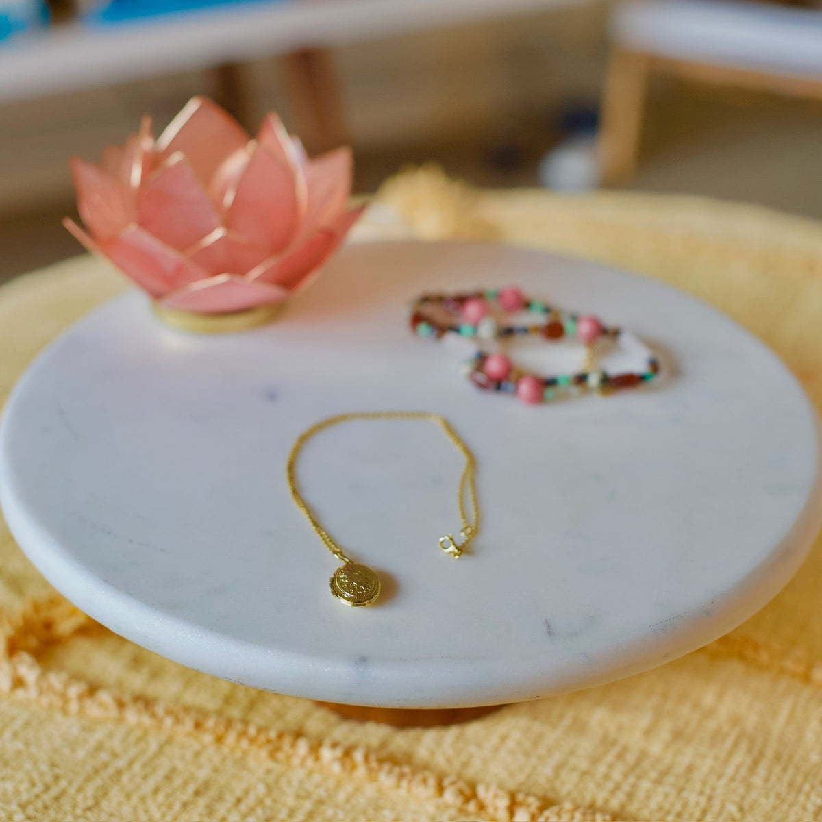 A gold-toned delicate bracelet with a small round disc charm and a star accent, displayed on a round white marble tray next to a pink lotus flower and a beaded bracelet on a yellow textured blanket.