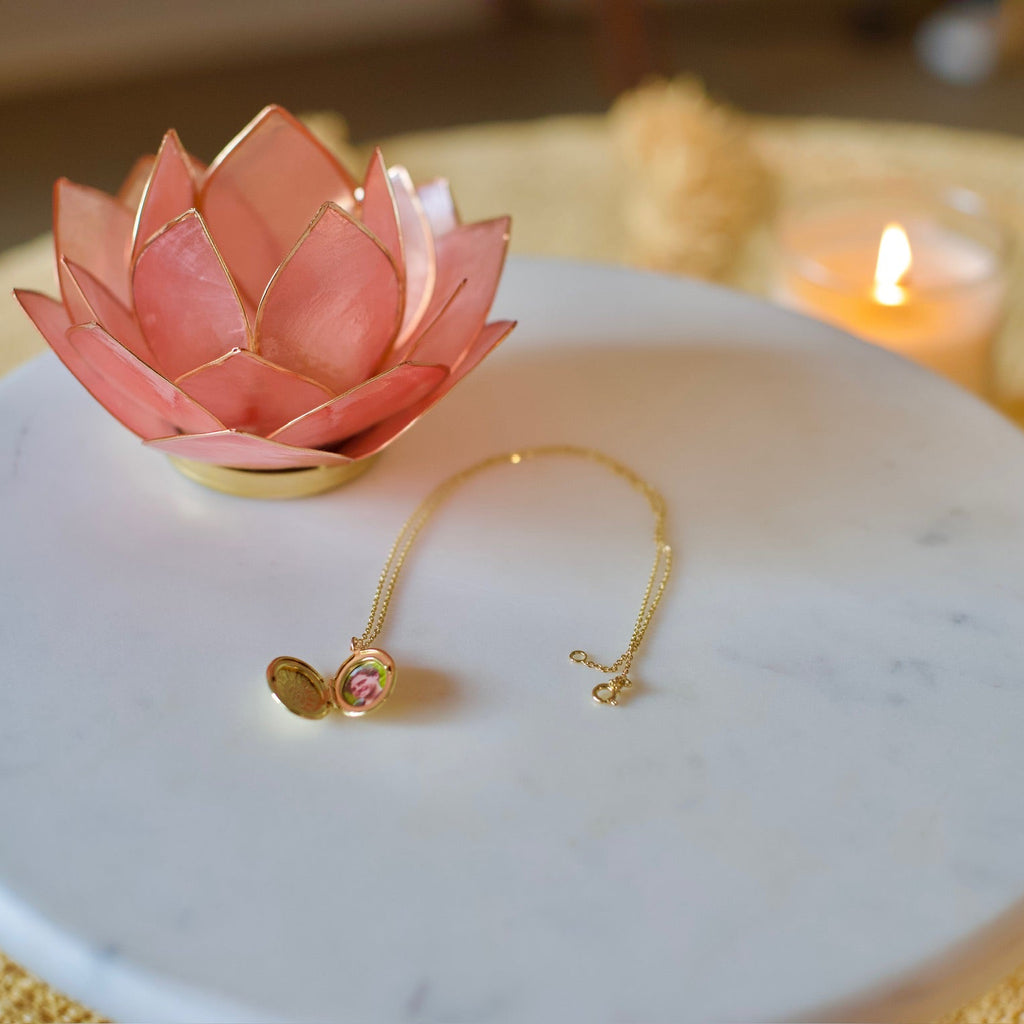 A gold-toned necklace with a small open heart pendant and a pink gemstone, displayed on a round white marble tray next to a pink lotus flower holder, on a yellow textured blanket with a blurred lit candle in the background.