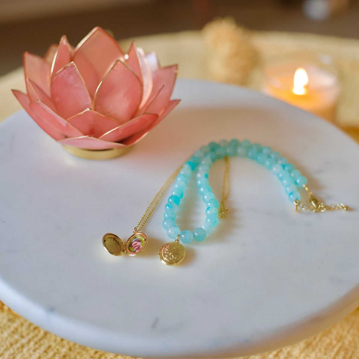 A gold-plated necklace with a pink gemstone and two gold charms, paired with a light blue beaded bracelet, displayed on a white round tray with a pink lotus candle holder and a lit candle in the background.