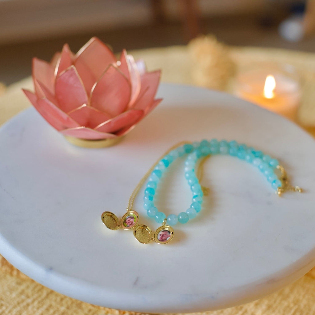 A beaded bracelet with light blue beads and gold-toned charms rests on a round white marble tray, styled with a pink lotus candle holder and a lit candle in a warm indoor setting.