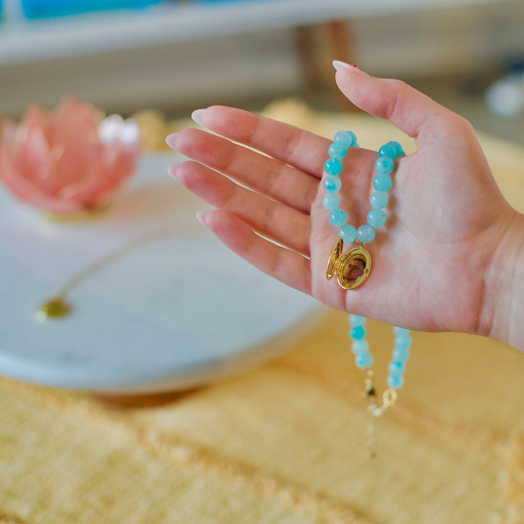 A hand holding a turquoise beaded necklace with a gold-toned zodiac sign pendant, set against a warm, softly blurred background.