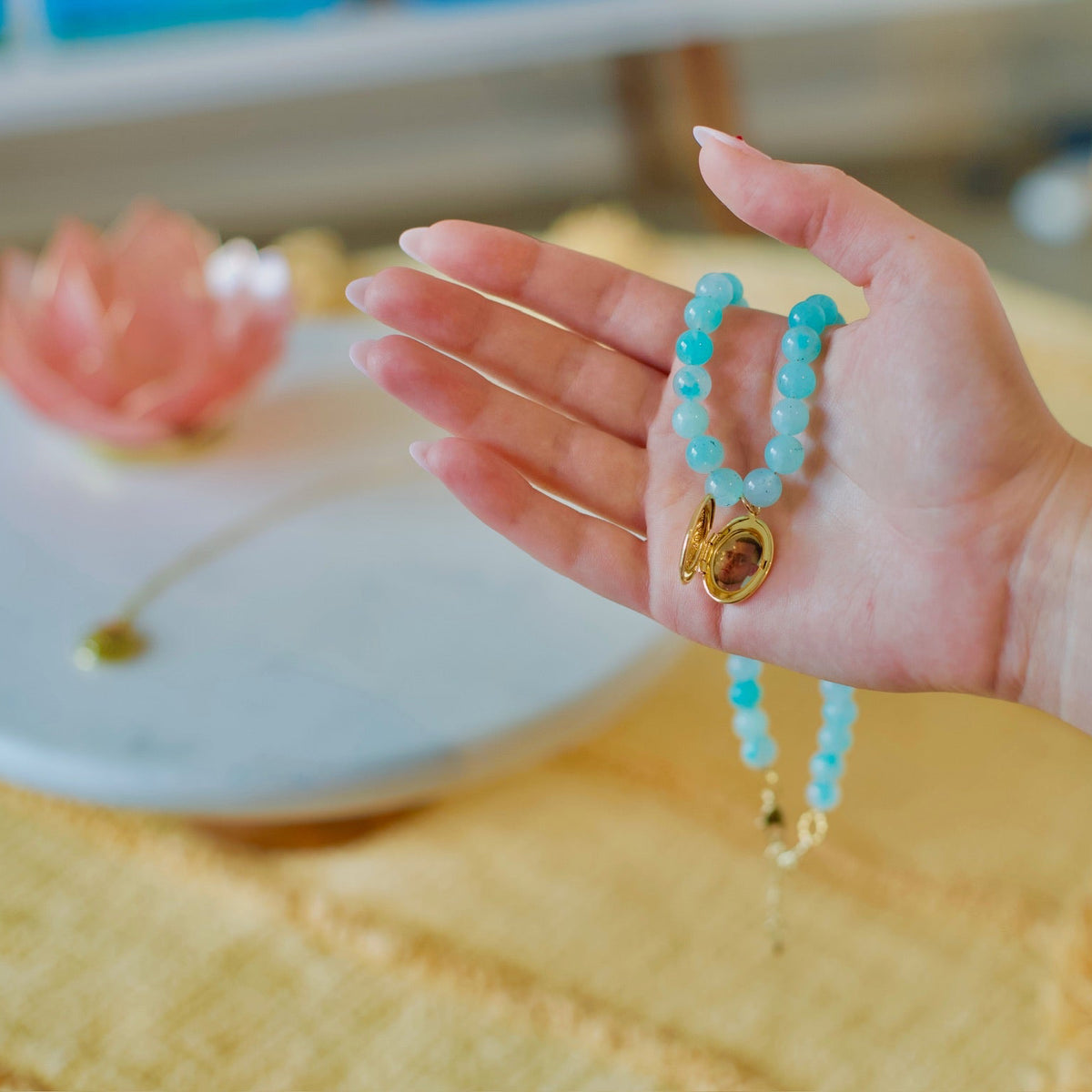 A hand holding a turquoise beaded necklace with a gold-toned zodiac sign pendant, set against a warm, softly blurred background.