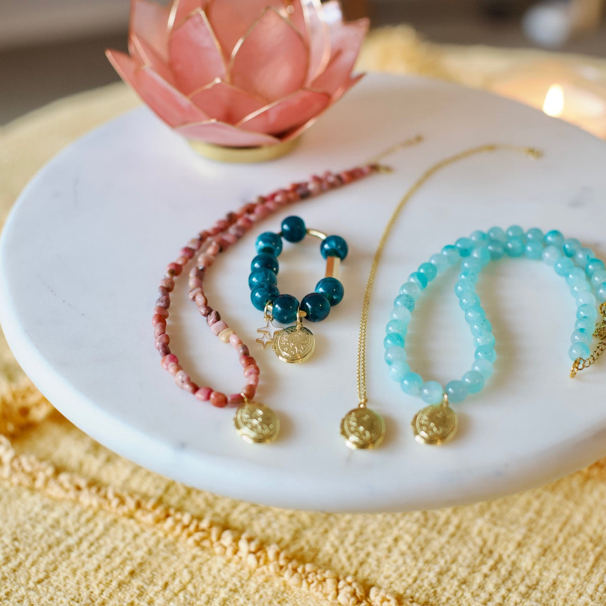 Three beaded bracelets with gold charms are arranged on a round white marble tray, set on a yellow textured blanket with a pink lotus flower and a blurred candle in the background.