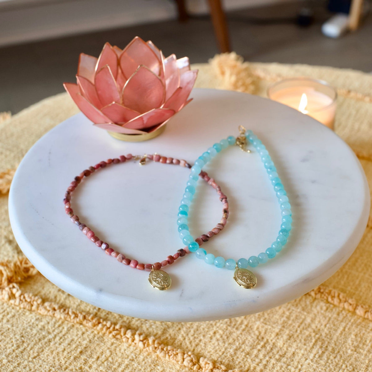 Two beaded bracelets, one in dusty pink and one in light blue, with gold coin charms, displayed on a white marble tray next to a pink lotus candle holder.