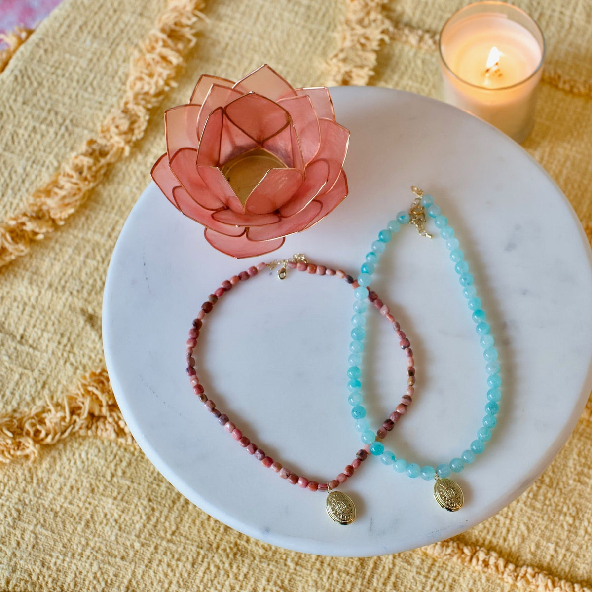 Two beaded bracelets, one in terracotta red and one in light blue, displayed on a round white marble plate alongside a pink lotus candle holder and a lit candle on a textured yellow fabric background.