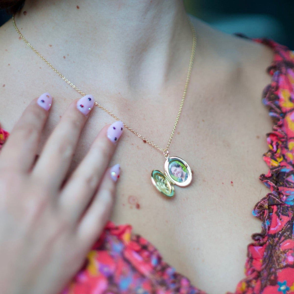 A close-up lifestyle shot of a woman wearing a delicate gold necklace with a double-oval pendant featuring green and pink mother-of-pearl inlay. She is wearing a vibrant, multicolored floral-patterned top, and her hand with pink glitter nail polish rests near her chest.