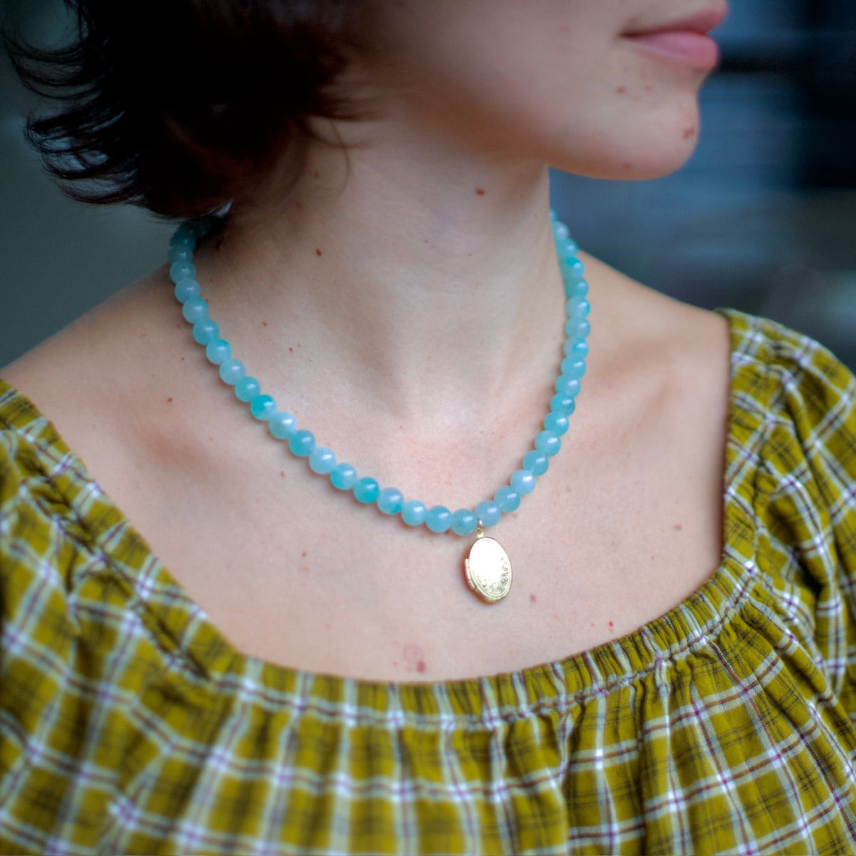 A close-up lifestyle shot of a woman wearing a light blue beaded necklace with a gold oval pendant, paired with a yellow and white plaid top.