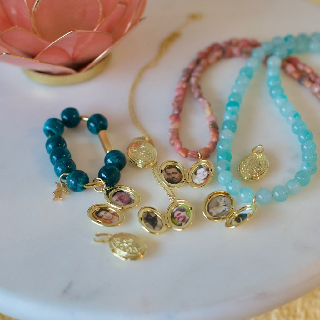 A styled flat-lay of several beaded jewelry pieces and gold charms arranged on a white marble tray, with a pink lotus candle holder and yellow textured fabric in the background.