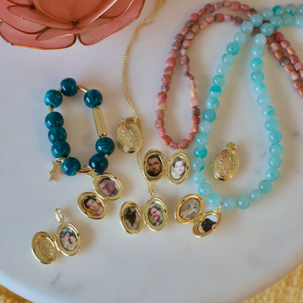 A styled flat-lay of assorted jewelry on a white round tray, including turquoise and pink beaded necklaces, a dark teal beaded bracelet, gold-tone locket pendants with portrait engravings, and a coral flower decoration on a yellow textured fabric background.