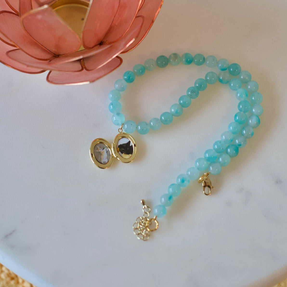 A turquoise beaded necklace with gold-toned locket and pendant charms, displayed on a round white marble tray next to a pink lotus candle holder.