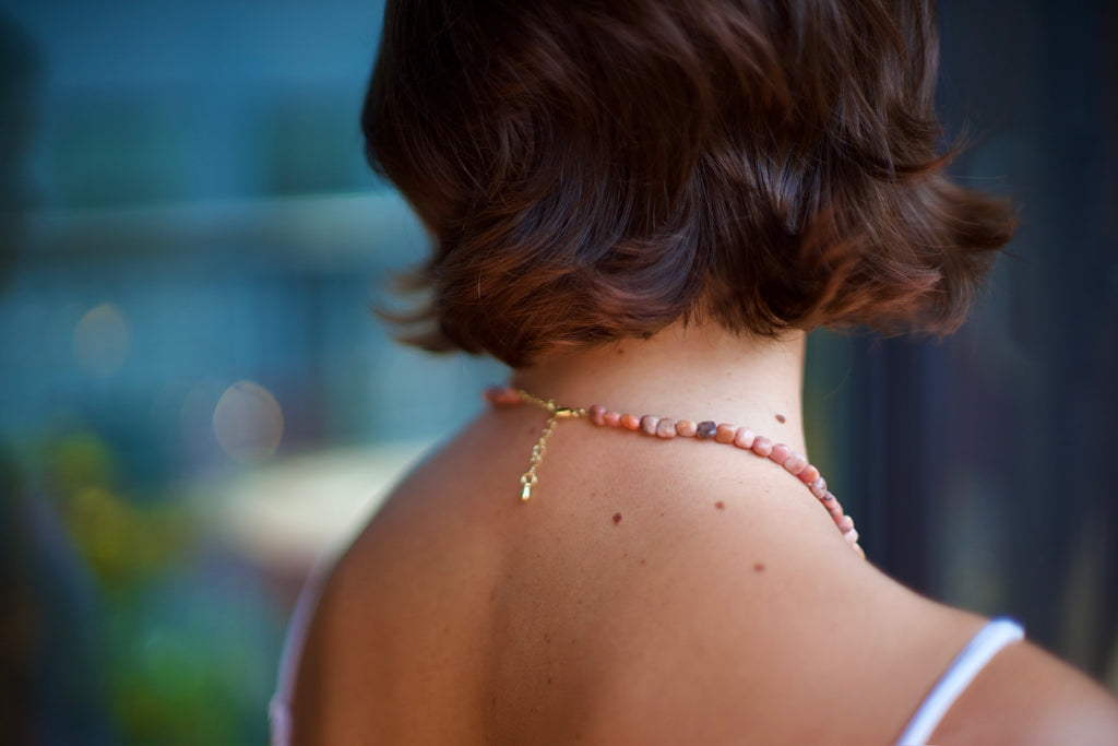 A close-up lifestyle shot of a woman from behind, showcasing a delicate pink beaded necklace with a gold clasp and charm worn around her neck.
