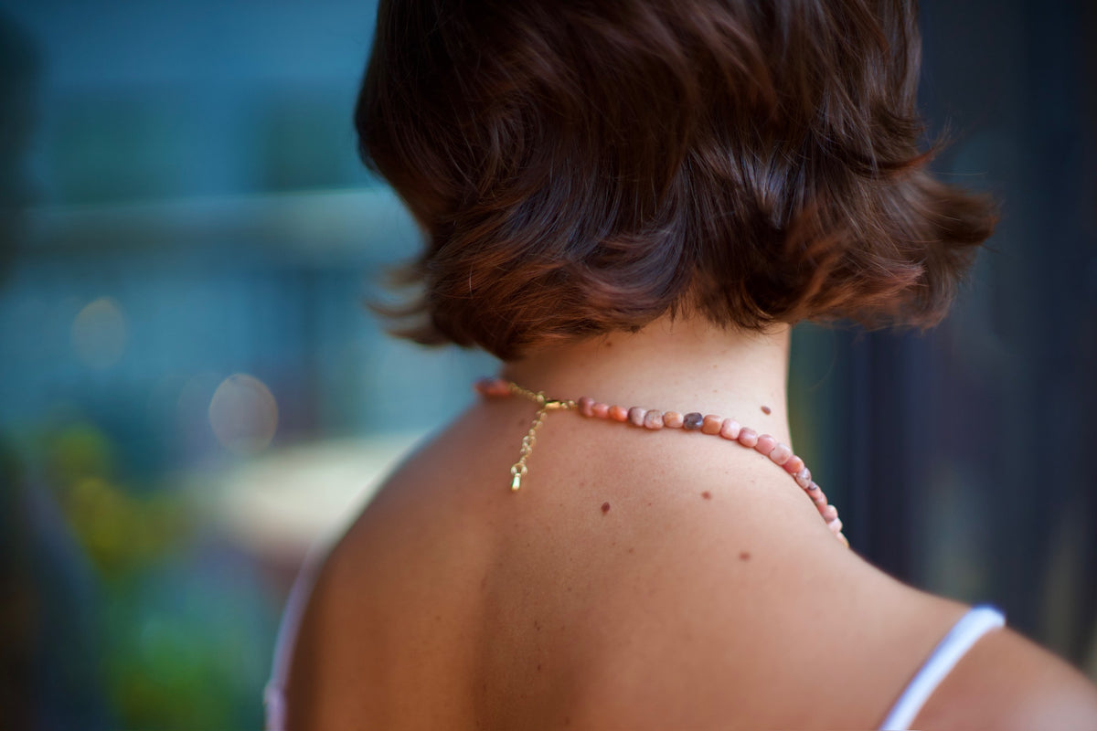 A close-up lifestyle shot of a woman from behind, showcasing a delicate pink beaded necklace with a gold clasp and charm worn around her neck.