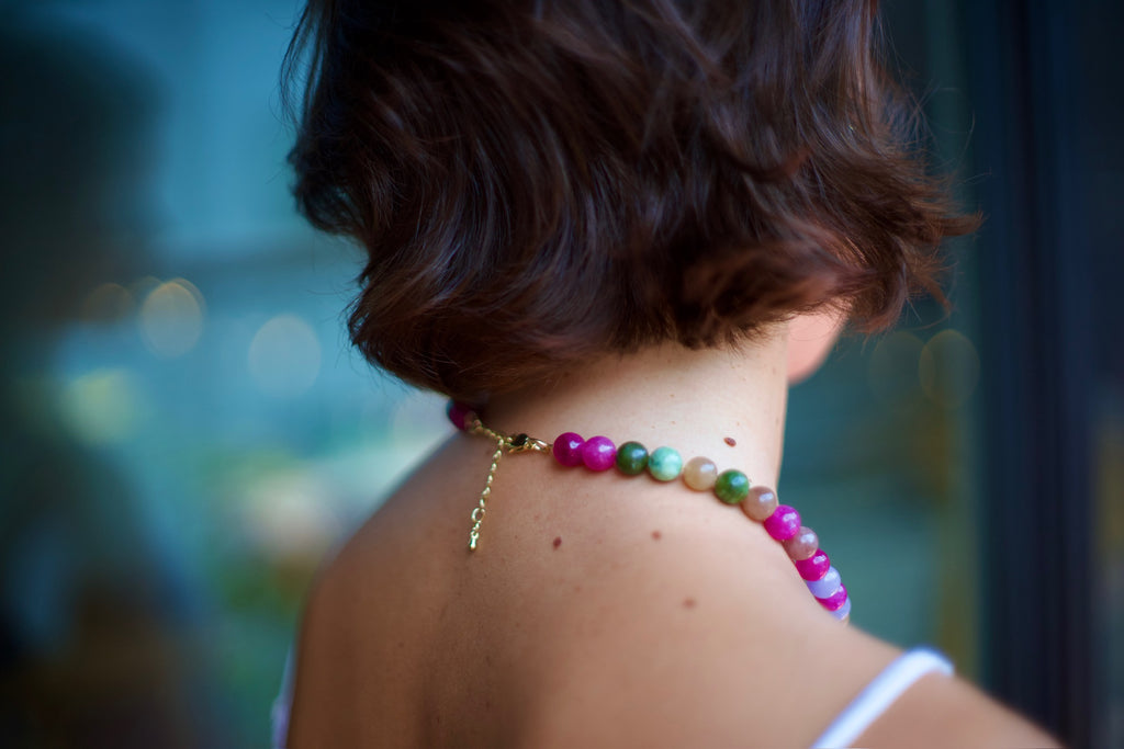 A close-up lifestyle shot of a woman from behind, showcasing a colorful beaded necklace with a gold clasp and chain. The necklace features a gradient of vibrant beads including pink, purple, green, and orange, worn against her neck.