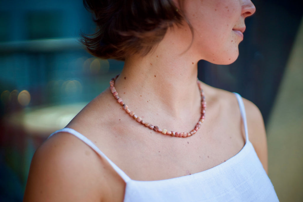 A close-up side profile of a woman wearing a delicate beaded necklace with terracotta, pink, and cream beads, paired with a white spaghetti-strap top.