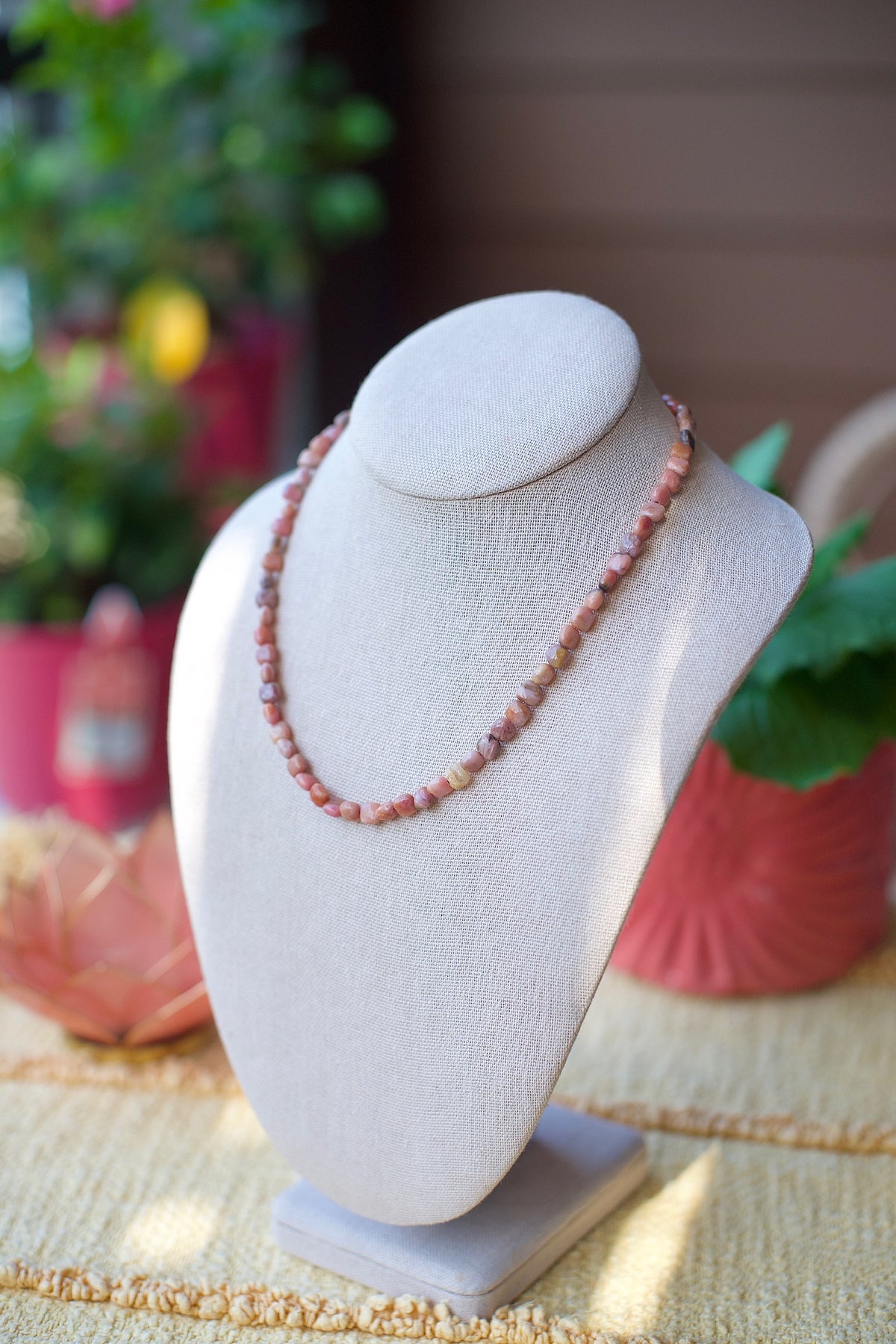 A beaded necklace made of pinkish-orange stone beads displayed on a light gray fabric jewelry bust, set against a softly blurred background with potted plants and warm wood tones.