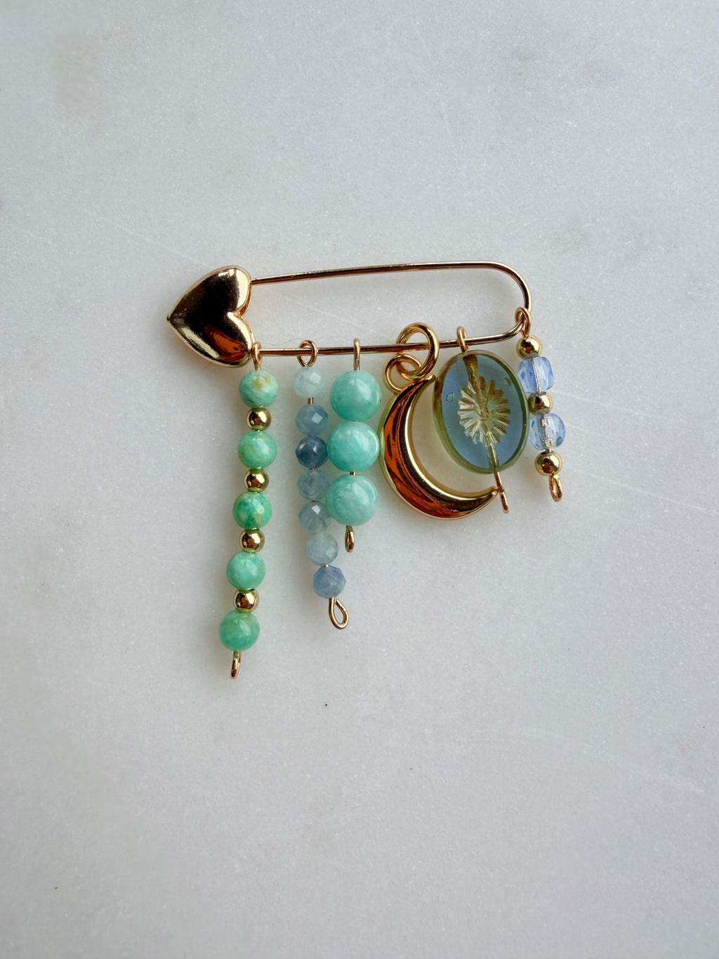 A gold-toned safety pin bag charm with dangling beaded charms, including mint green beads, light blue beads, a crescent moon charm, and a round sun-face medallion, displayed on a light gray background.