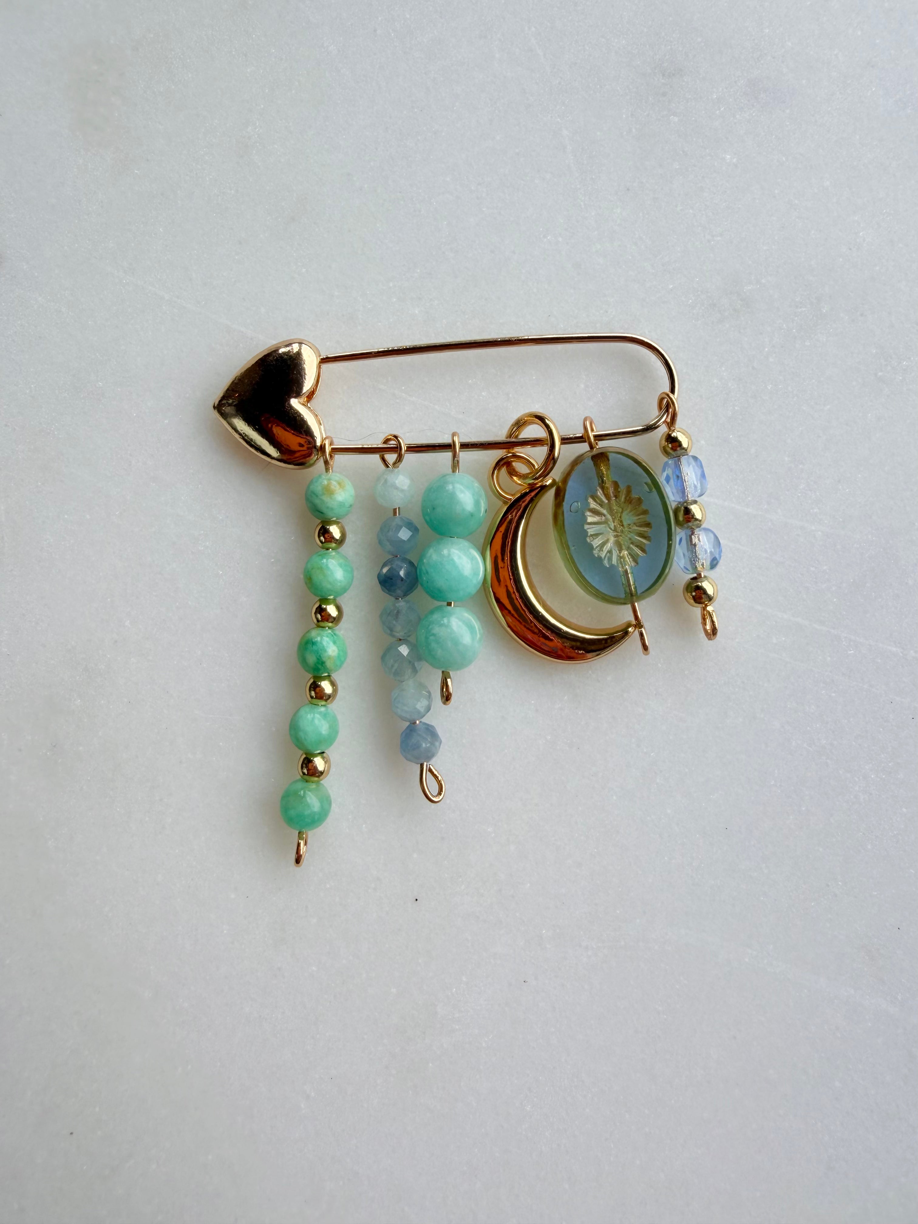 A gold-toned safety pin bag charm with dangling beaded charms, including mint green beads, light blue beads, a crescent moon charm, and a round sun-face medallion, displayed on a light gray background.