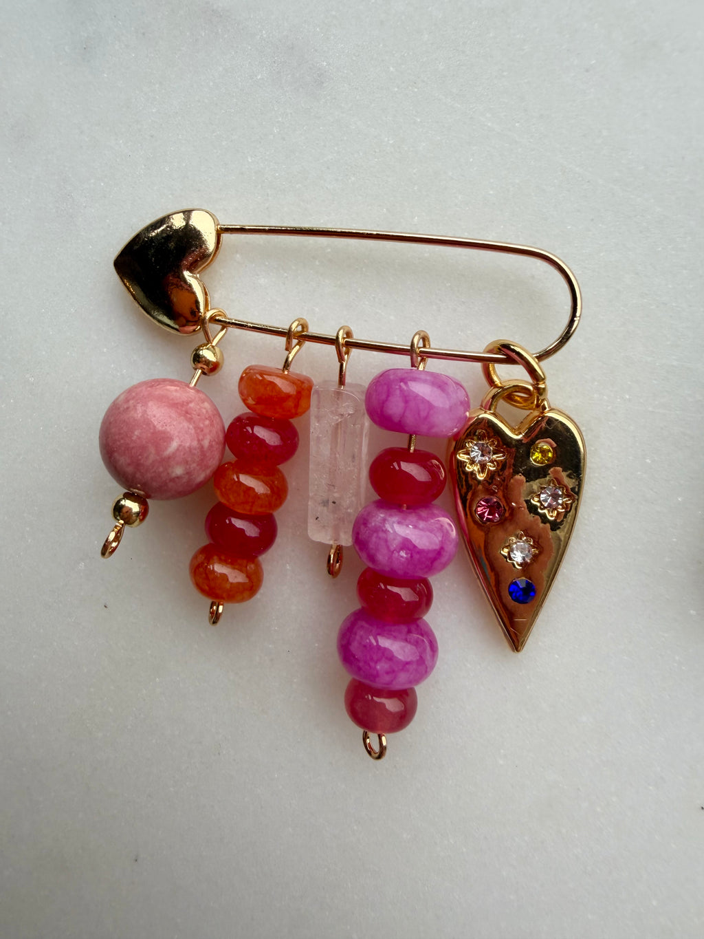 A gold-tone safety pin bag charm decorated with dangling heart-shaped charms, including pink and orange beads, a clear crystal, and a heart-shaped charm embellished with colorful rhinestones, shown on a light gray background.
