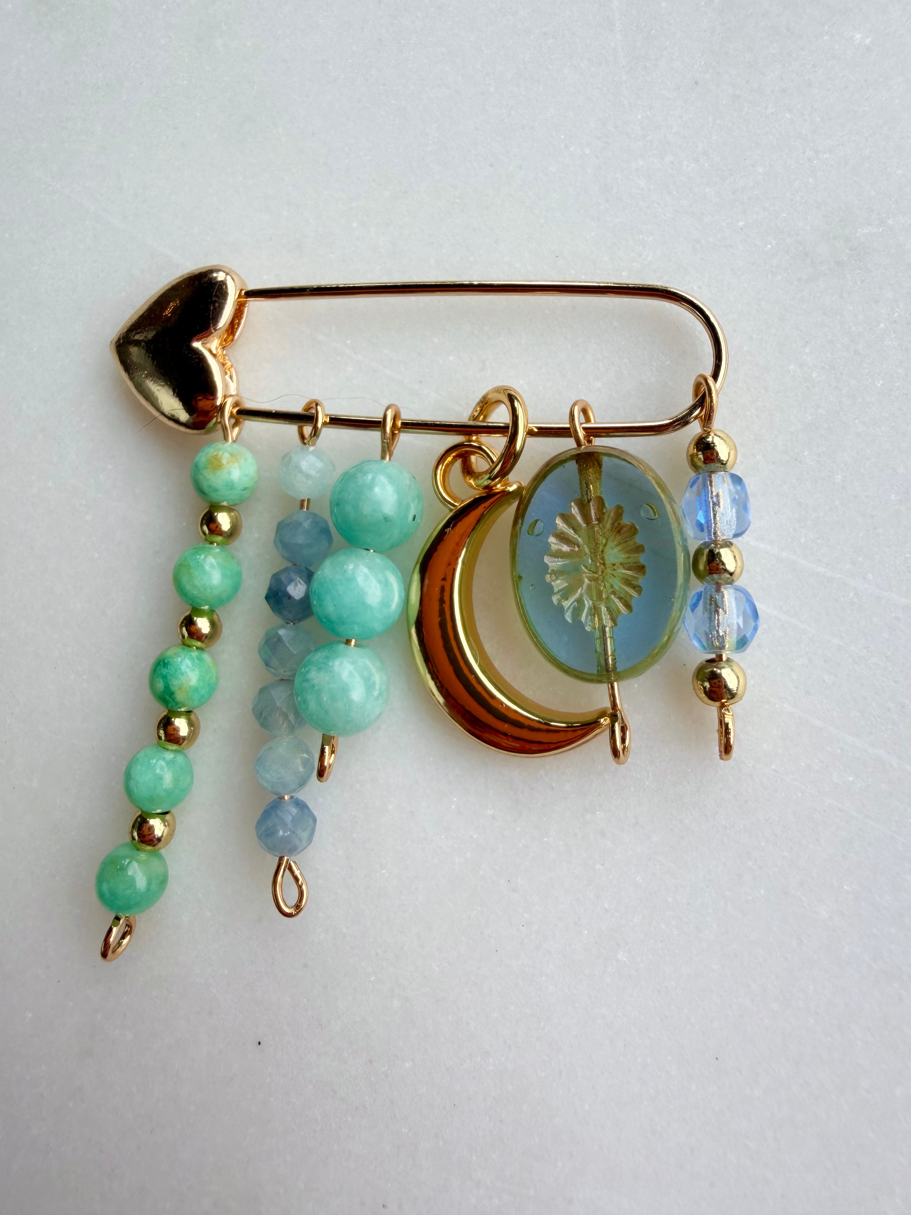 A gold-toned safety pin bag charm decorated with dangling charms, including a gold heart, green and blue beads, a crescent moon with a sun motif, and faceted beads, shown on a light gray background.