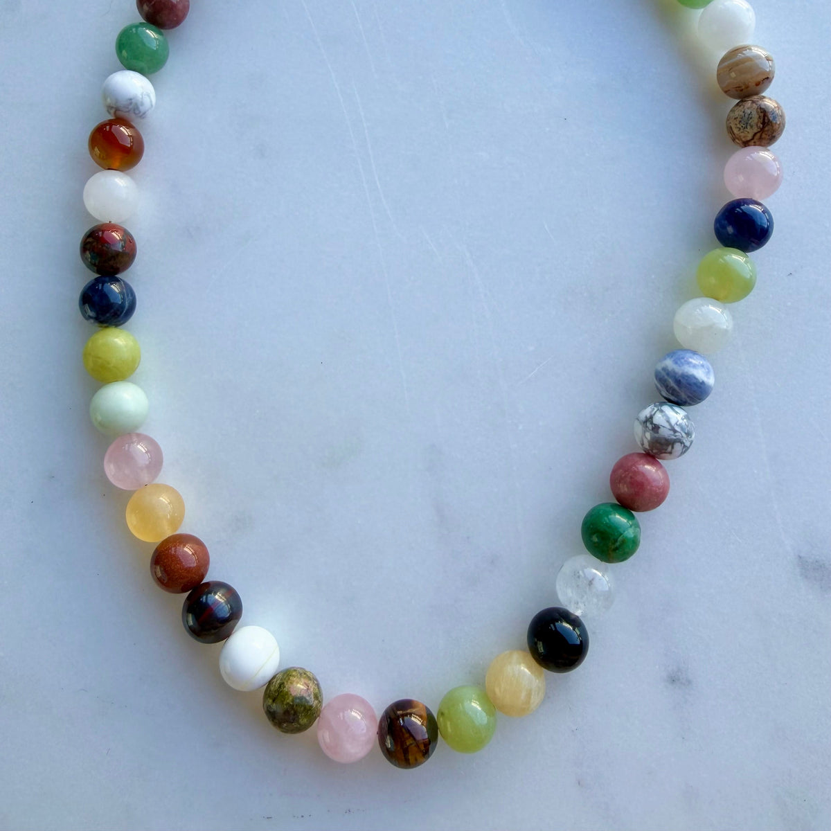 A beaded necklace made of polished multicolored gemstone-like beads, arranged in an oval shape on a light gray marble surface. The necklace features a gold-tone clasp and extender chain.