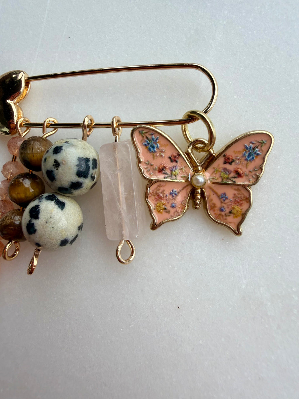 A gold-tone safety pin brooch decorated with a pink butterfly charm, blue floral details, a clear quartz crystal, and two white beads with black spots, shown on a light background.