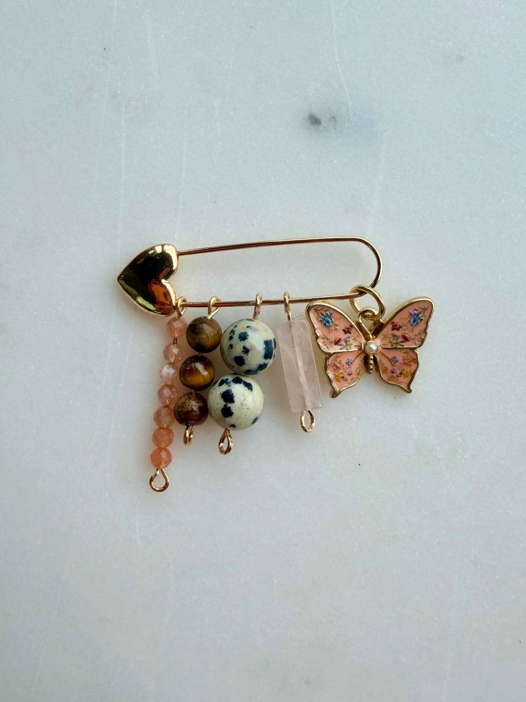 A gold-toned safety pin bag charm decorated with a pink and blue enamel butterfly, white and brown spotted beads, a pink beaded drop, and a clear quartz crystal, displayed on a light gray surface.