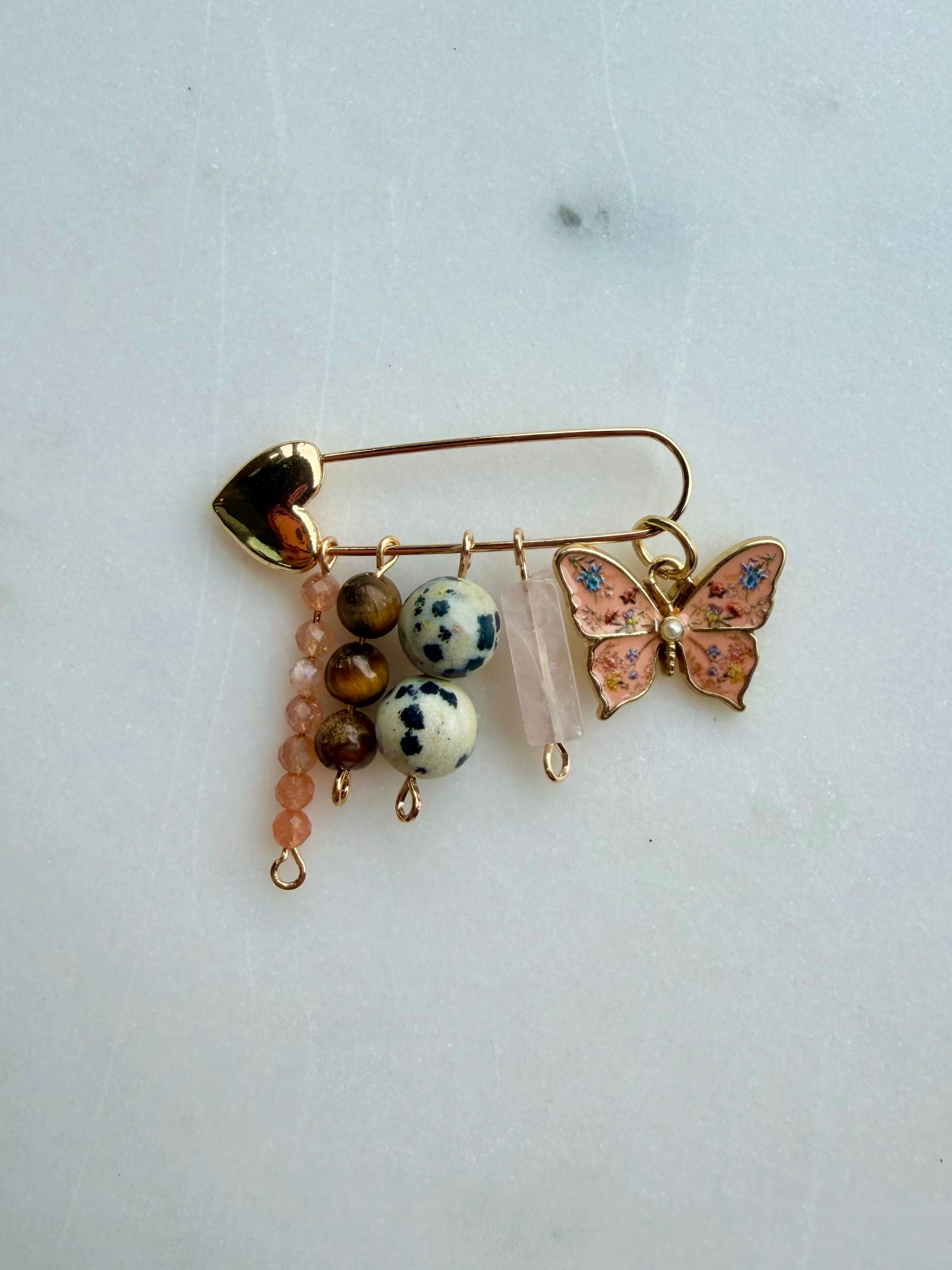 A gold-toned safety pin bag charm decorated with a pink and blue enamel butterfly, white and brown spotted beads, a pink beaded drop, and a clear quartz crystal, displayed on a light gray surface.