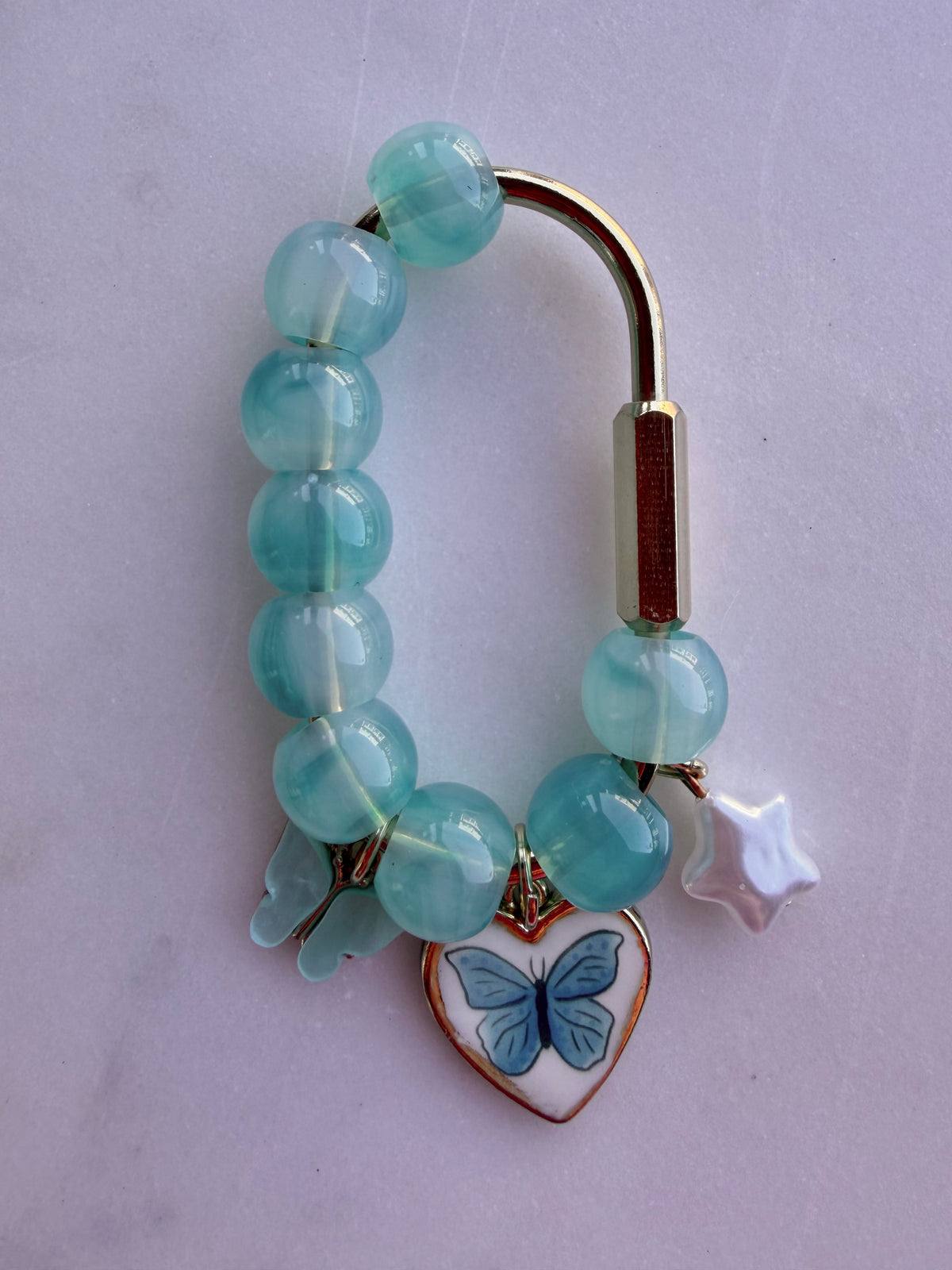 A gold bag charm features blue beads with a white star, blue butterfly heart charm, and a blue butterfly